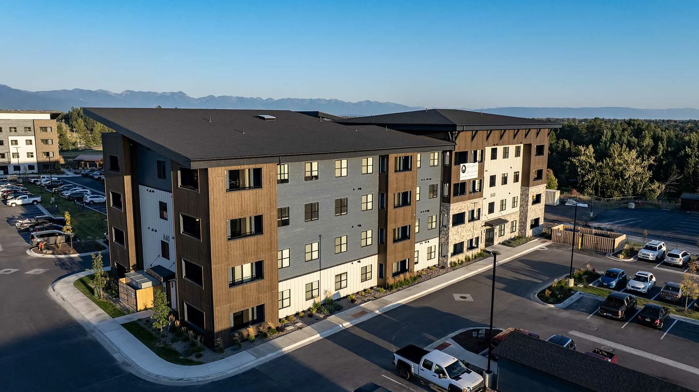 Modern apartment complex surrounded by lush greenery and scenic mountain views, offering ample parking and stylish exterior design.