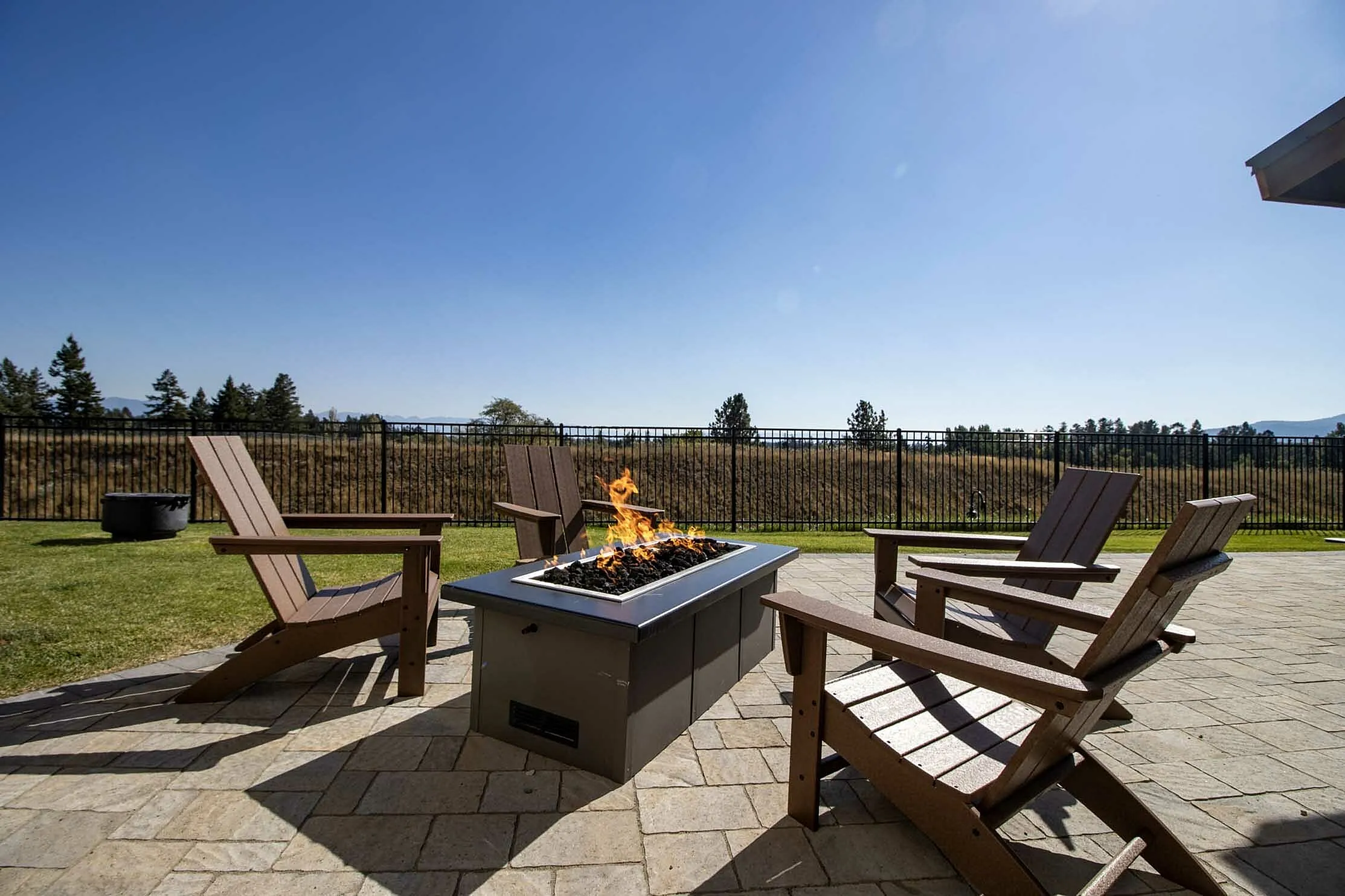 Inviting outdoor patio with Adirondack chairs surrounding a modern fire pit, offering scenic views of an open field and blue sky.