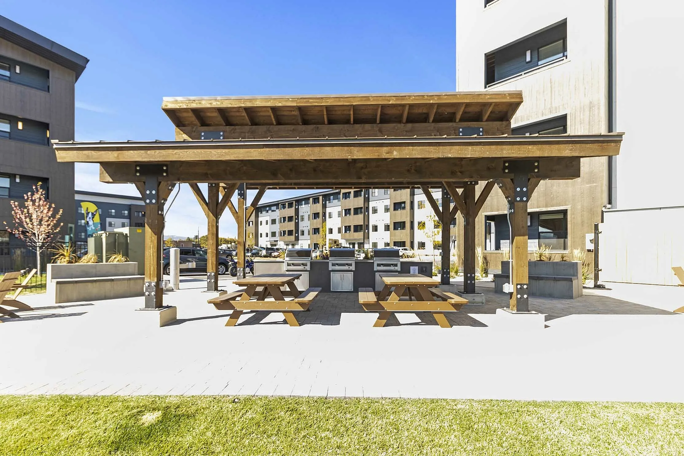 Modern outdoor patio area with wooden pergola, picnic tables, and built-in grills, surrounded by stylish apartment buildings.
