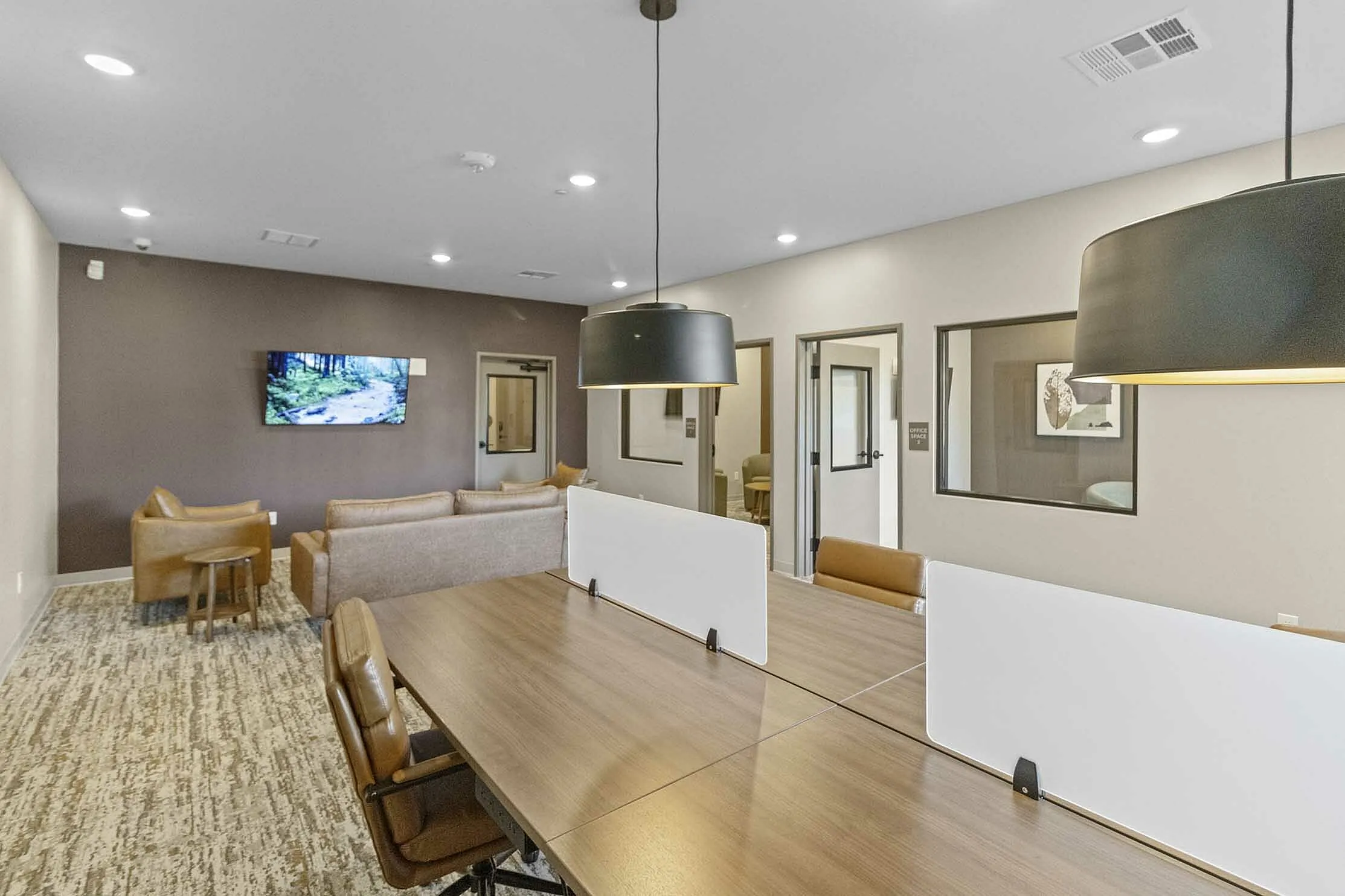 Modern coworking space with stylish furniture, featuring leather chairs, a large wooden table with dividers, and a cozy lounge area with a wall-mounted TV.