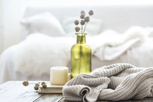 Cozy bedroom scene with a green vase, soft knits, and ambient candle on a rustic wooden table, inviting relaxation.