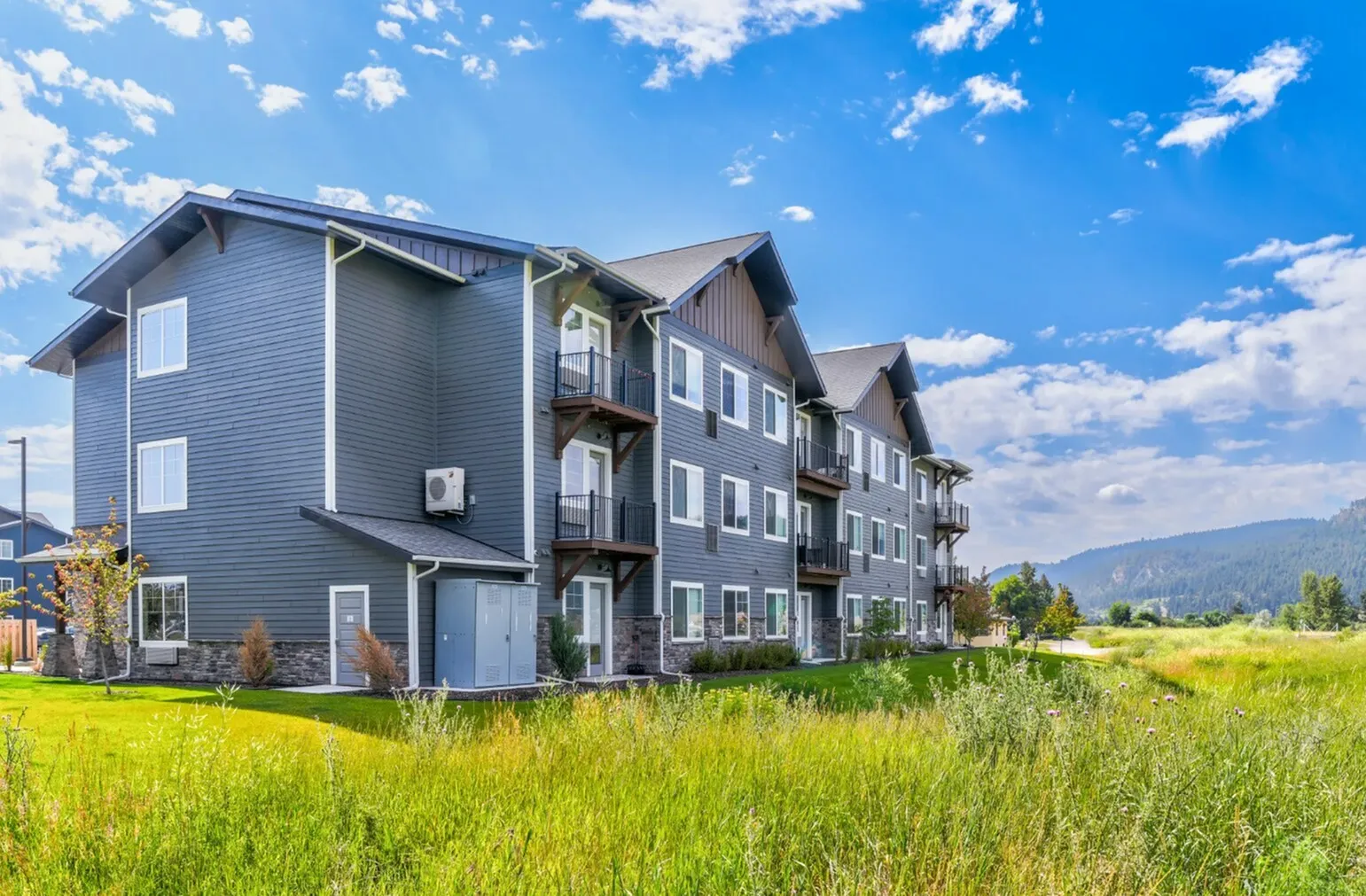 Modern three-story apartment building with balconies, nestled in lush greenery and scenic mountain views under a vibrant blue sky.