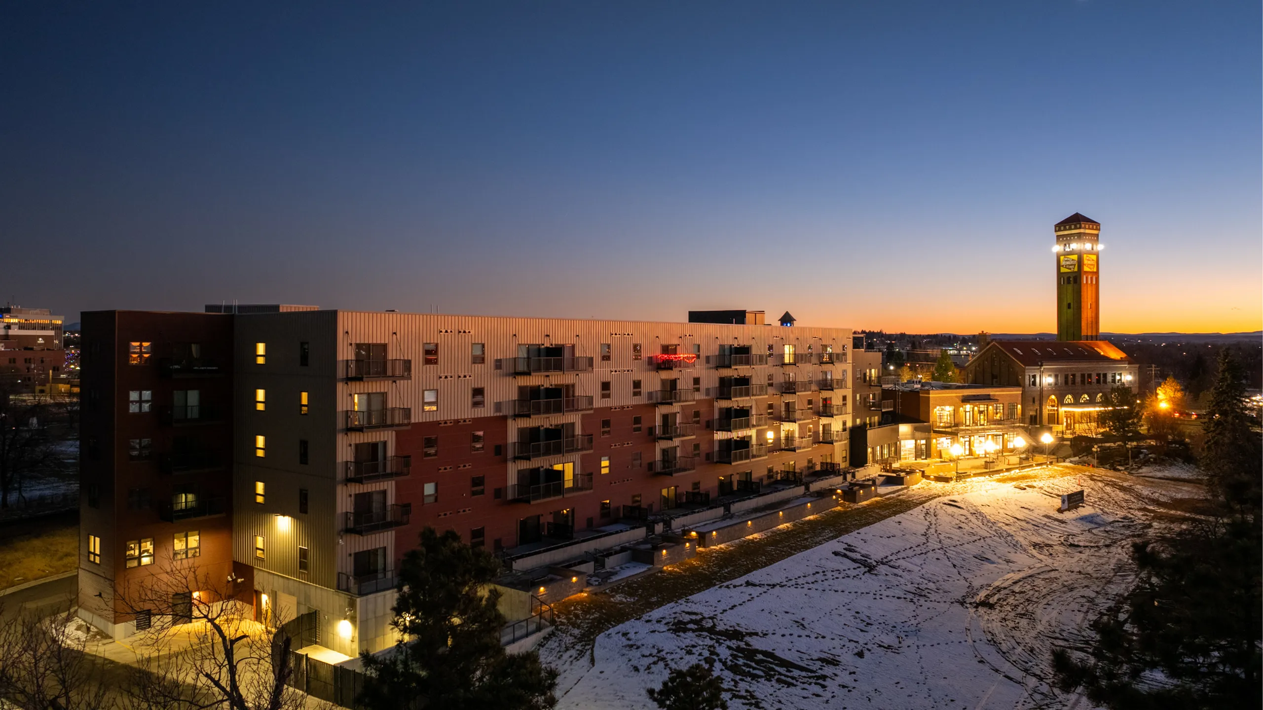 Chic urban apartment buildings with illuminated windows at dusk, showcasing a snowy courtyard and a distant tower.