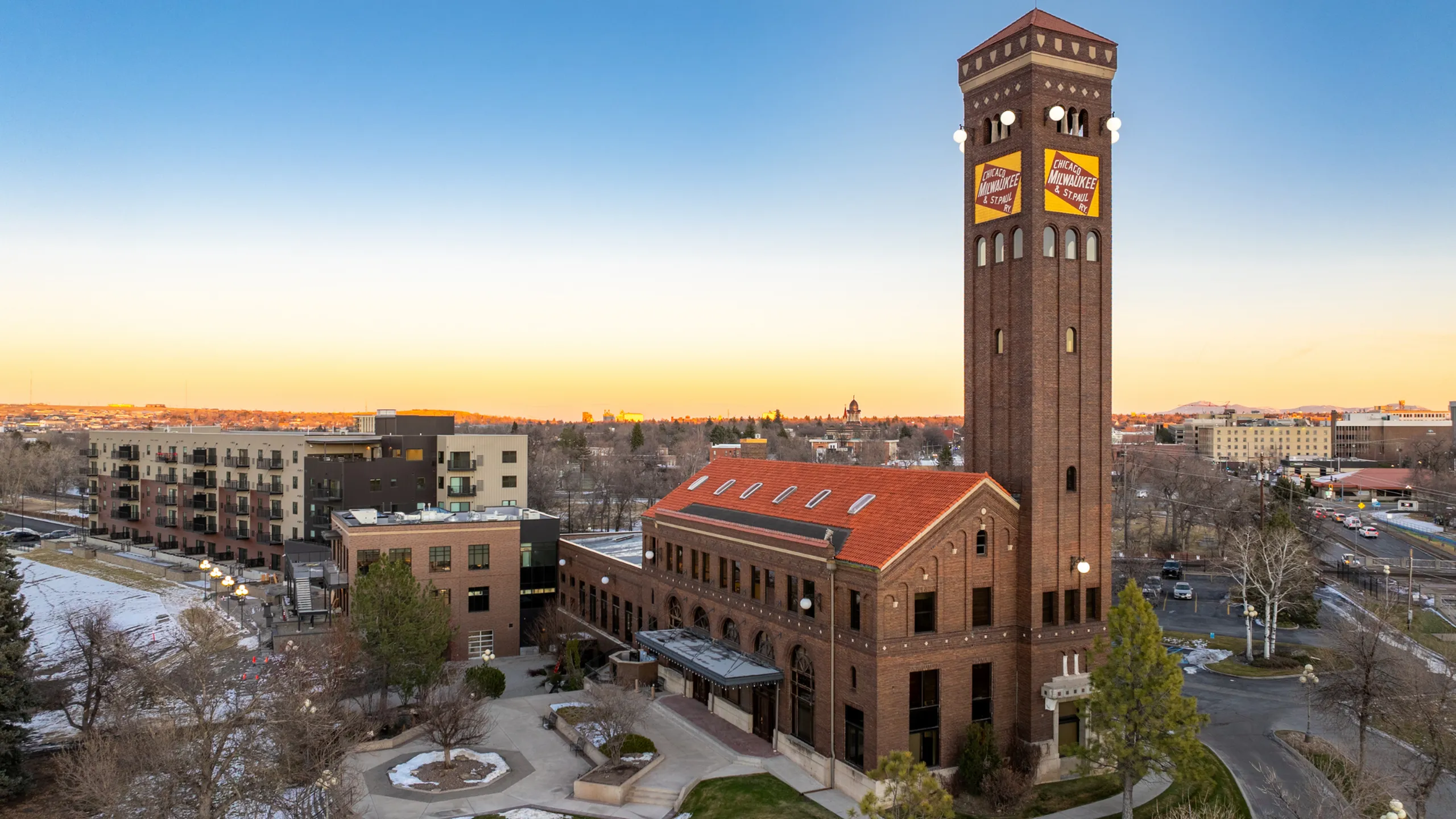Stunning brick building with a tall tower at sunset, surrounded by modern apartments and cityscape.