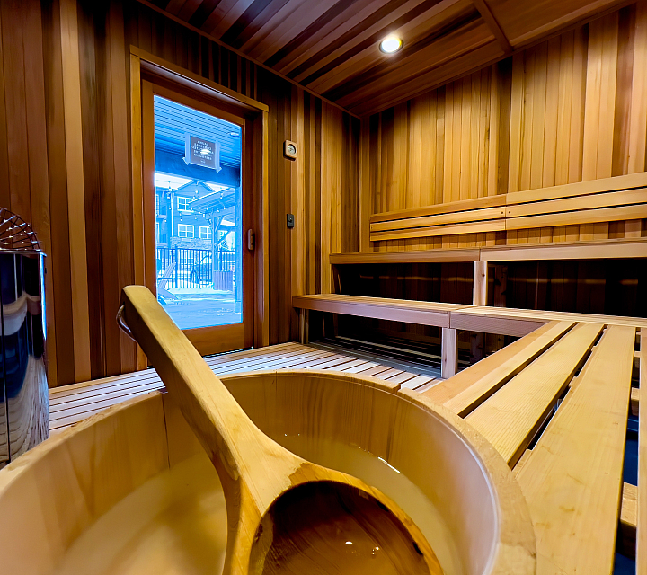 Elegant wooden sauna room with seating and a large sauna bucket, offering relaxation with a view through the glass door.