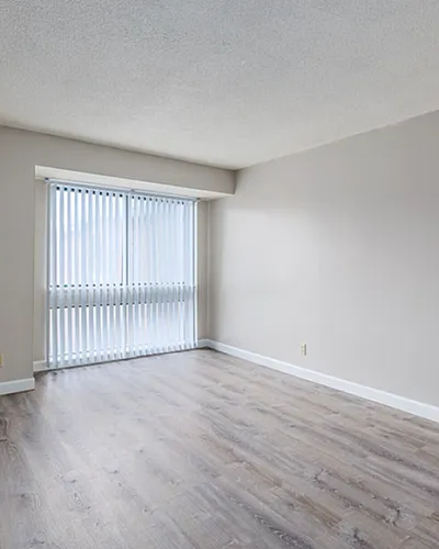 Bright living area showcasing modern wood flooring, large window with vertical blinds, and a glimpse of a tiled kitchen with warm cabinetry.