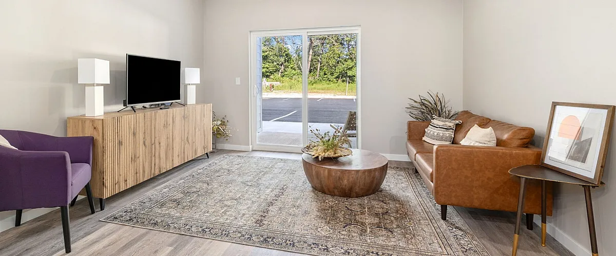 Modern living room with stylish decor, featuring a sleek brown sofa, elegant wooden furniture, and a plush area rug. Large windows offer natural light and serene outdoor views.