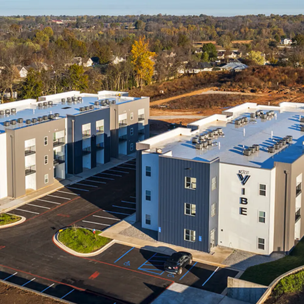 Aerial view of modern apartment complex with sleek design, ample parking, and surrounded by lush greenery, offering tranquil living in a vibrant neighborhood.