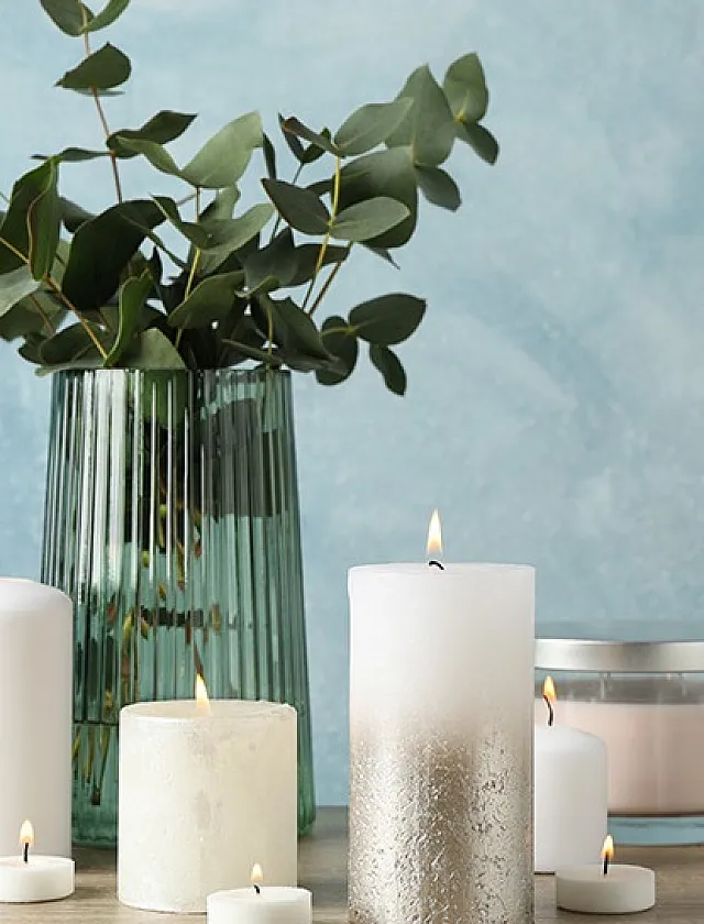 Elegant arrangement of white candles and green foliage in a glass vase against a textured blue backdrop.