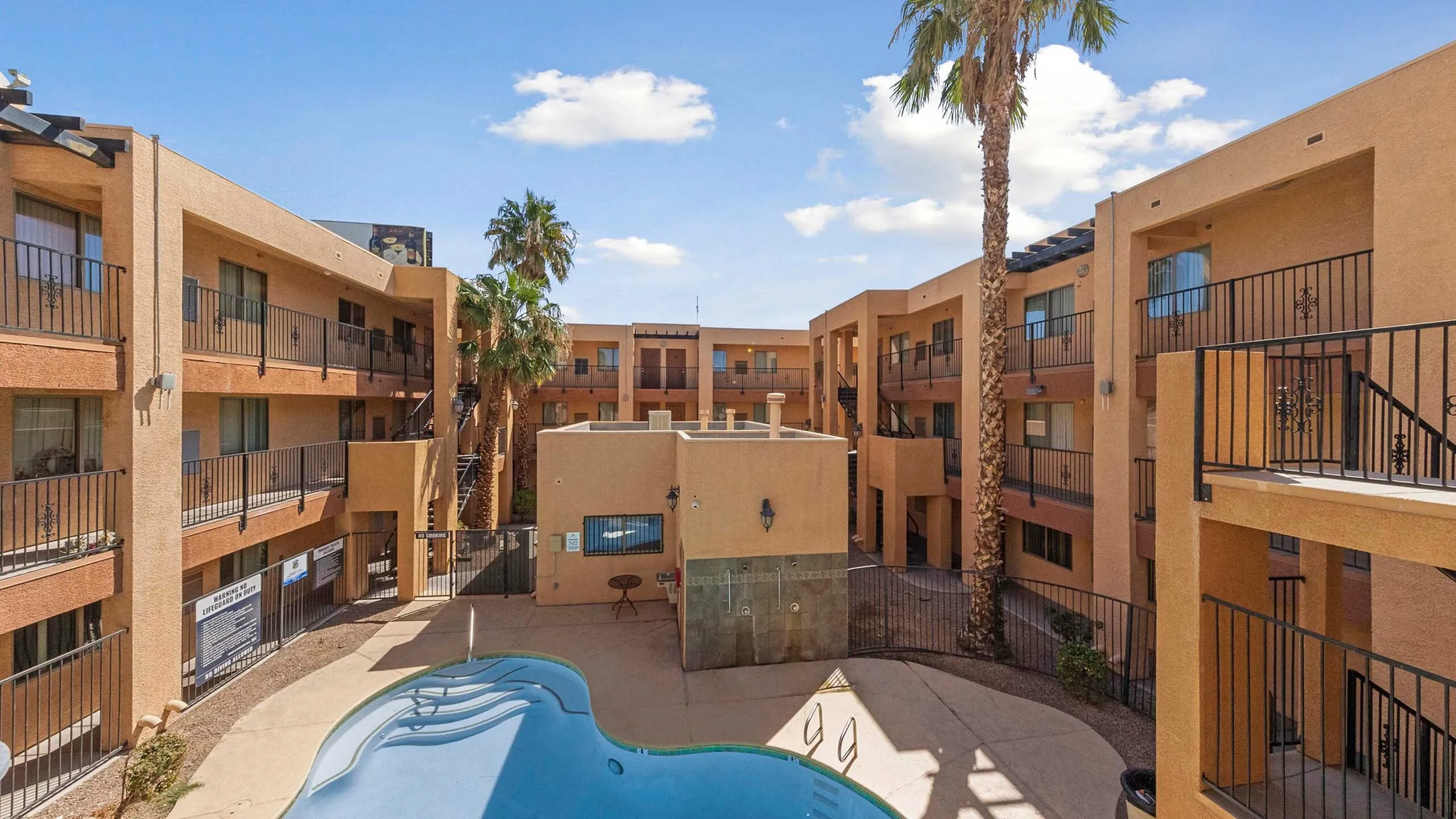 Spacious apartment courtyard with a pool and palm trees under a bright blue sky.