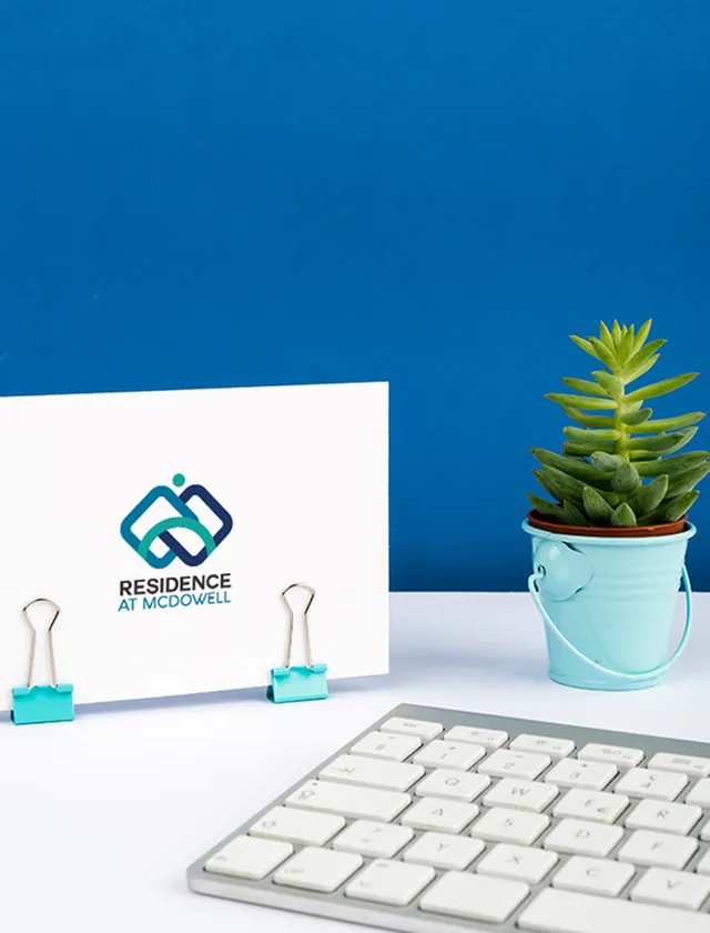 Stylish desk setup featuring a 'Residence at McDowell' card held by turquoise clips, a vibrant potted succulent, and a sleek white keyboard against a bold blue backdrop.