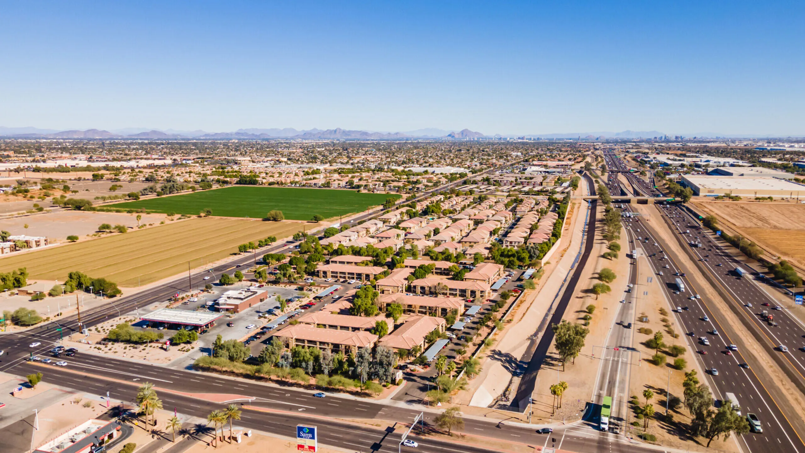 Scenic aerial view of a spacious suburban neighborhood featuring neatly arranged homes with terracotta roofs surrounded by lush greenery, adjacent to major highways, and set against a backdrop of distant mountain ranges.