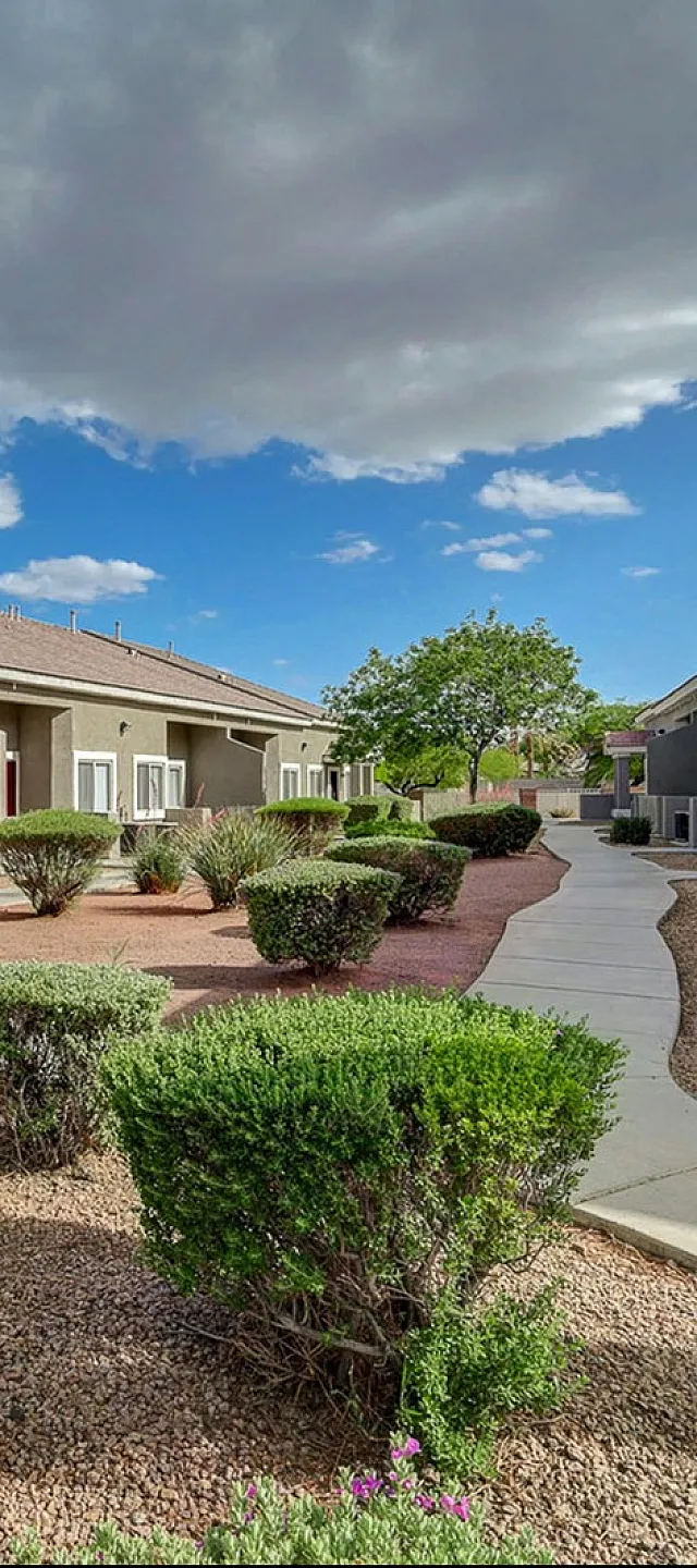 Charming apartment complex with neatly landscaped pathways, vibrant green shrubs, and a bright blue sky backdrop.