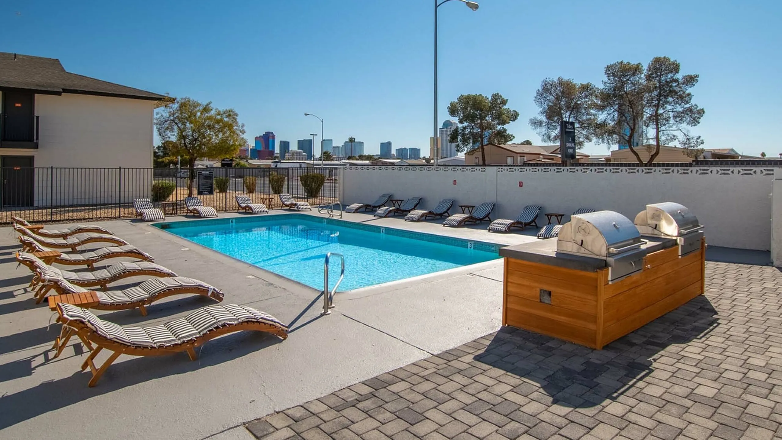 Chic apartment complex featuring a pristine swimming pool, elegant sun loungers, and modern BBQ grills, set against a backdrop of city skyline and lush greenery.