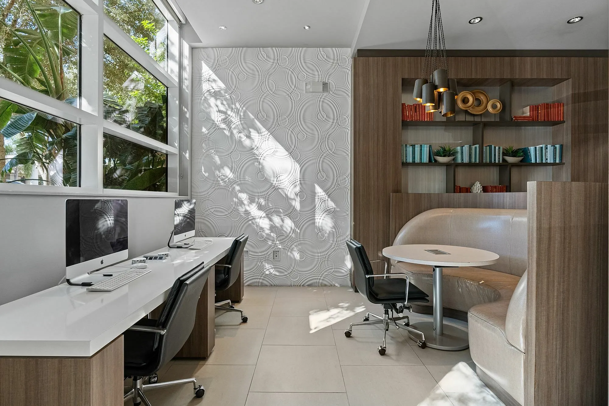 Contemporary workspace featuring sleek computer stations, cozy seating, and a stylish bookshelf under ample natural light.