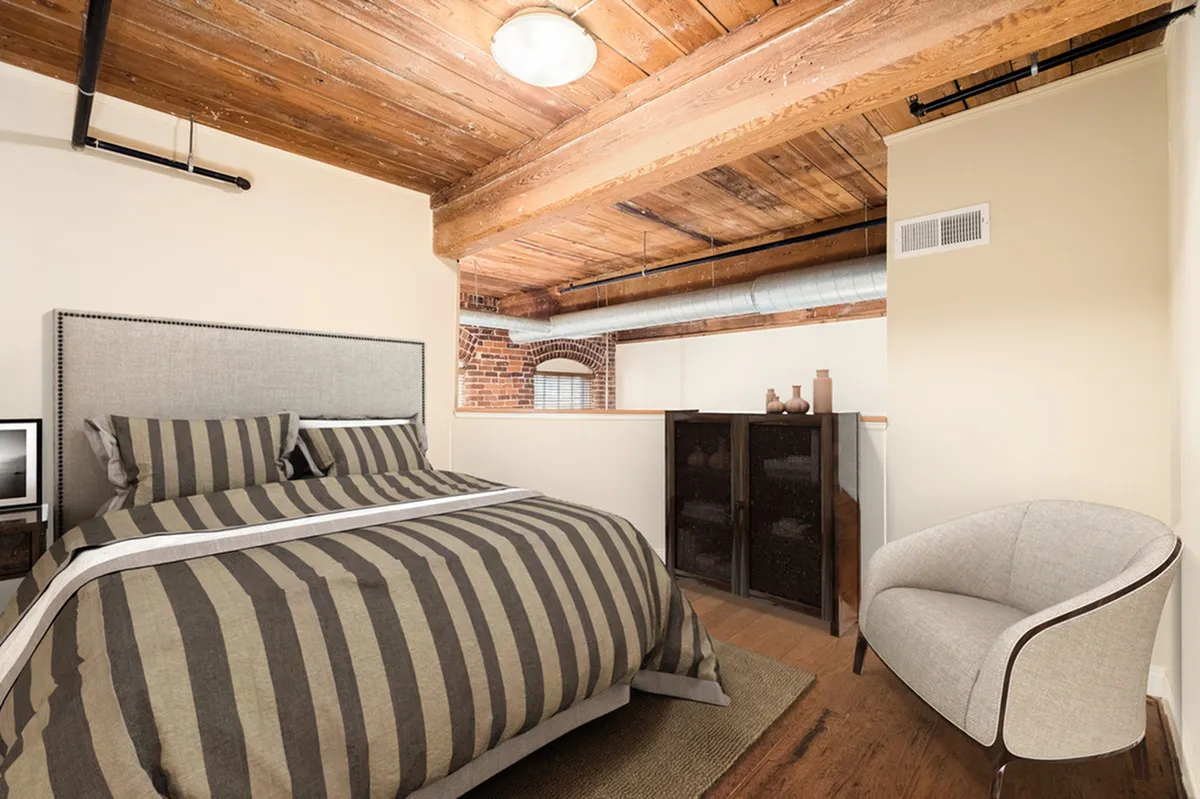 Chic and cozy bedroom at Alpha Mill Apartments in Charlotte, NC with exposed wooden ceiling, featuring a comfortable striped bed and modern decor.