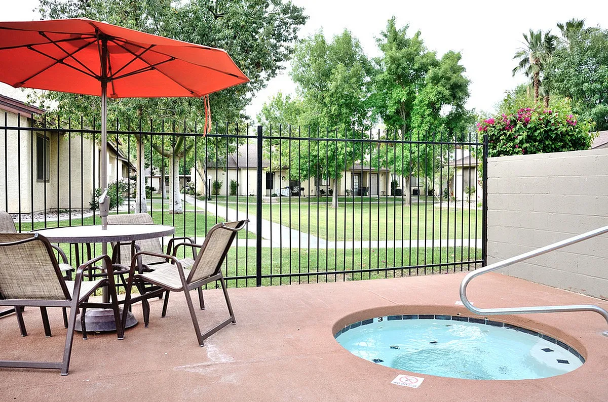 Charming outdoor patio at Camelot in Yuma with cozy seating, vibrant red umbrella, and inviting hot tub, overlooking lush green garden.