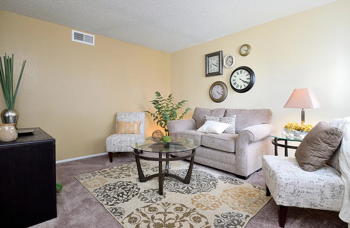 Inviting living room with plush seating, stylish decor, and a glass coffee table, accentuated by charming wall clocks and lush plants at Camelot Apartments in Arizona.