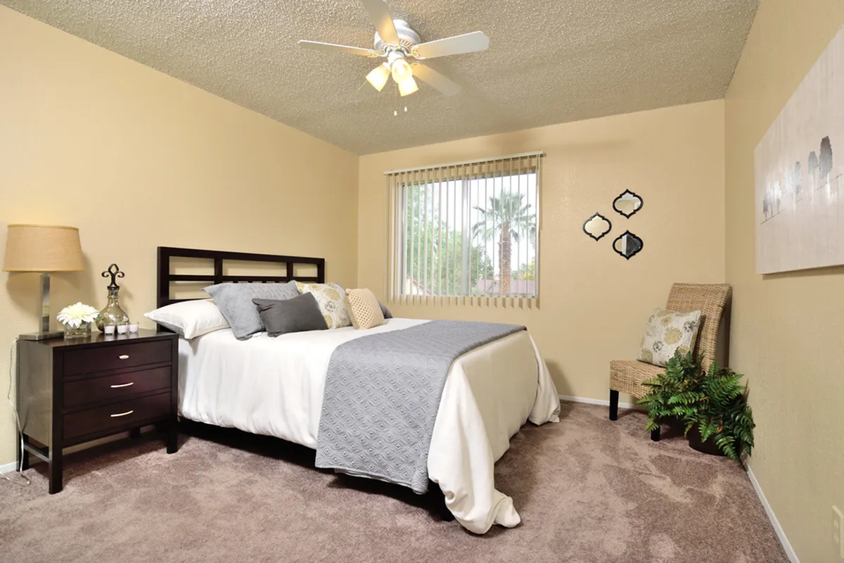 Camelot Apartment's inviting bedroom with plush carpet, elegant decor, and cozy bed. Natural light filters through vertical blinds, enhancing the serene ambiance.