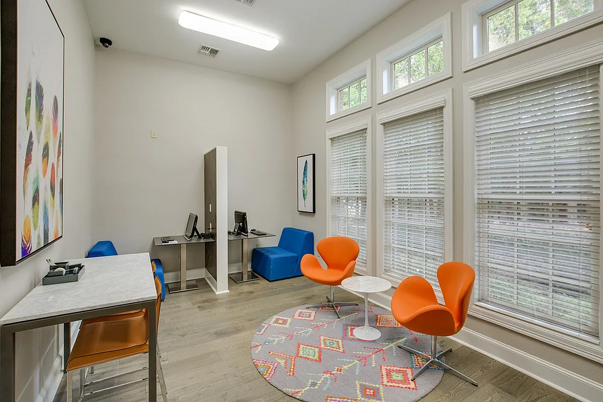 Raveneaux Apartments' contemporary lounge area featuring vibrant orange chairs, a sleek work station, and ample natural light from large windows.