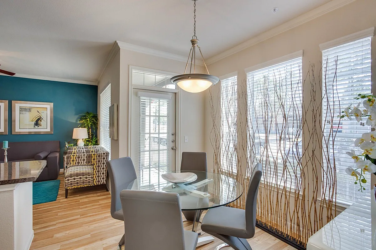 Bright and inviting dining area with a modern glass table, elegant chairs, and abundant natural light streaming through large windows with decorative blinds at Raveneaux Apartments.