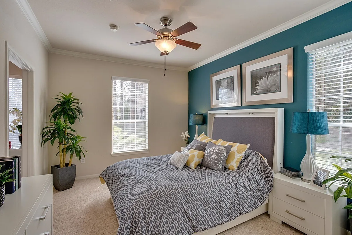 Inviting model bedroom at Raveneaux Apartments with a modern design, featuring a cozy bed adorned with patterned pillows, vivid teal accent wall, and stylish decor elements. Abundant natural light streams through large windows, creating a serene and welcoming atmosphere.