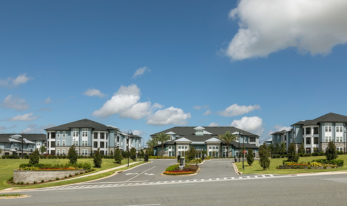 Entrance of Cottonwood Clermont Apartments surrounded by lush gardens and a clear blue sky, offering serene living and picturesque views.