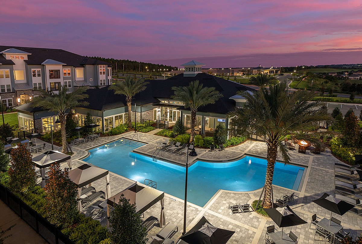 Aerial view of Cottonwood Clermont Apartments featuring a stunning illuminated pool area with palm trees and elegant lounge chairs, set against a breathtaking sunset backdrop.