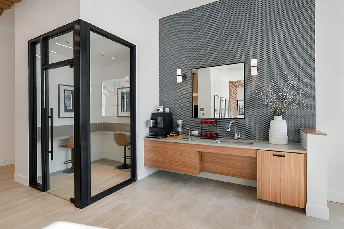 View of Apt CoWork at Cottonwood Highland featuring a sleek glass office pod, elegant wooden cabinetry with a coffee station, and stylish decor with a statement wall mirror.