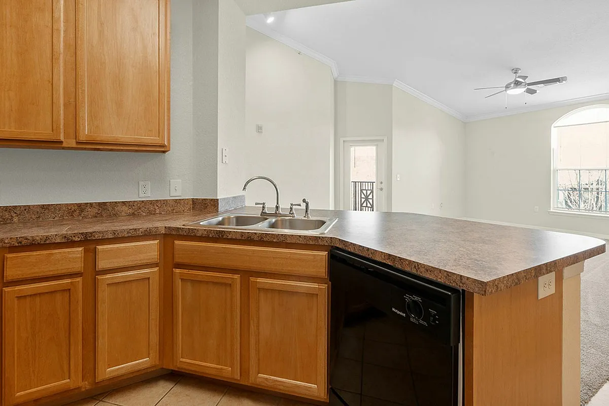 Bright and inviting kitchen at Cottonwood Reserve Apartments with elegant wooden cabinetry, modern double sink, and spacious countertop, leading into an open-concept living area with abundant natural light.