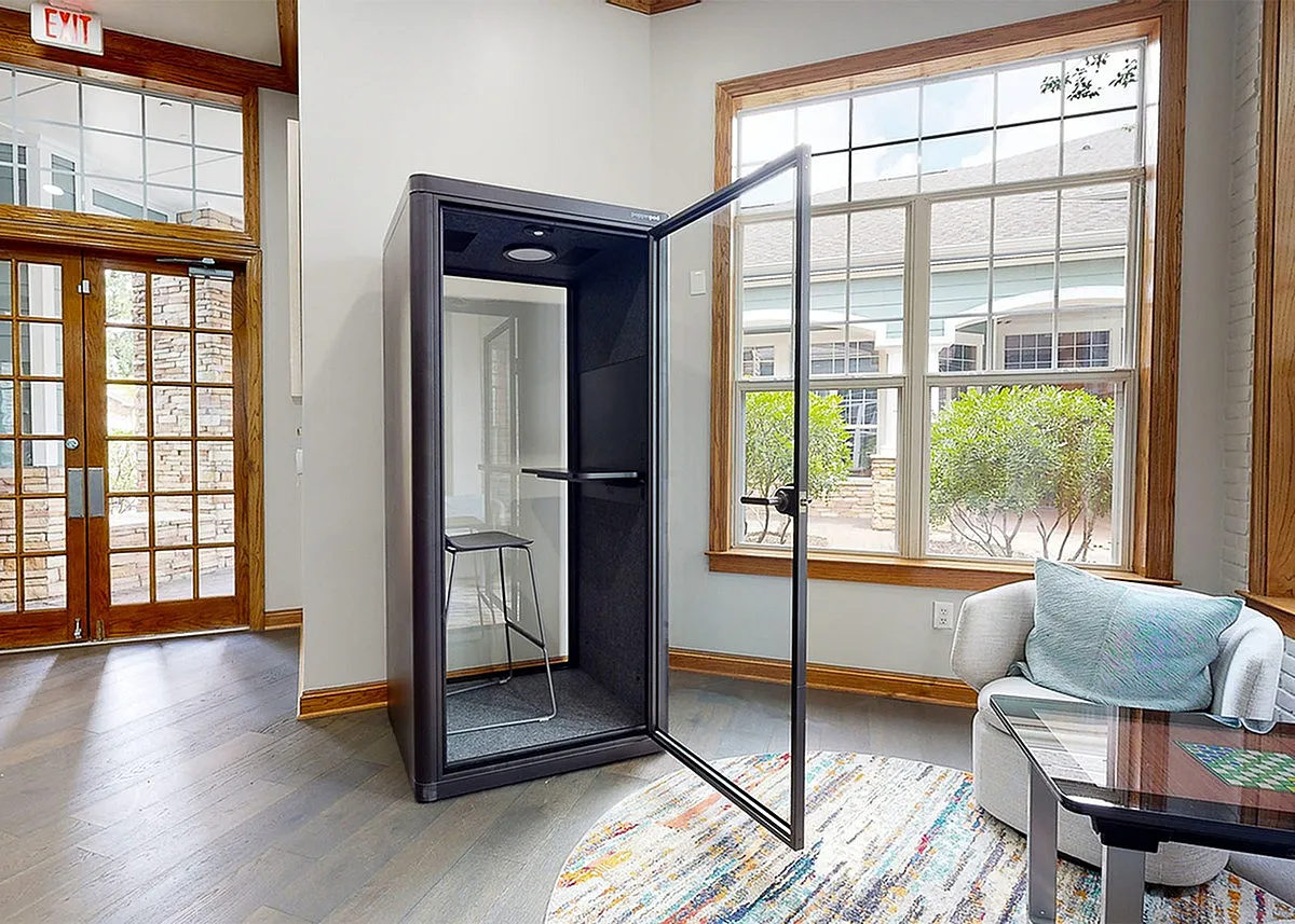 Modern office space at Cottonwood Ridgeview Apartments featuring a sleek phone booth with glass door, showcasing a tranquil view through large windows and elegant wooden accents.