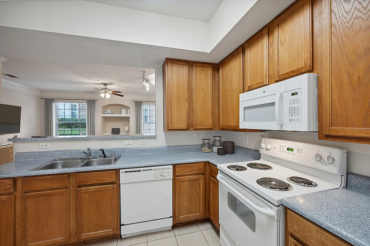 Modern kitchen at Cottonwood Ridgeview Apartments with sleek countertops, wooden cabinets, and built-in appliances, opening to a bright living area with large windows.
