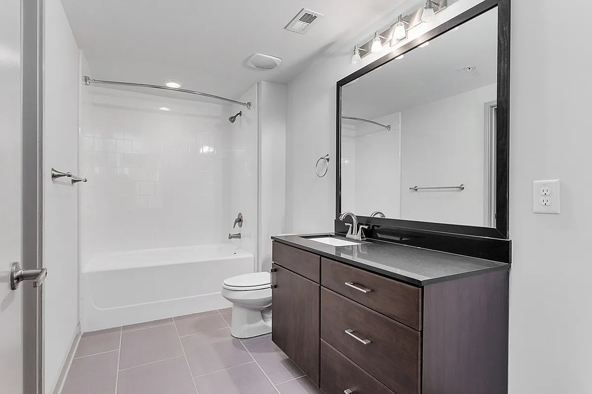 Modern bathroom at Cottonwood Westside Apartments featuring a sleek dark wood vanity, spacious mirror, and elegant fixtures, complemented by a pristine white bathtub and polished tile flooring.