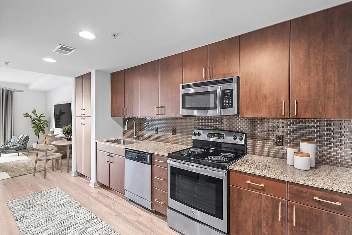 Modern kitchen at Cottonwood Westside Apartments with stainless steel appliances and elegant wood cabinetry, leading to a cozy living area with stylish furniture.