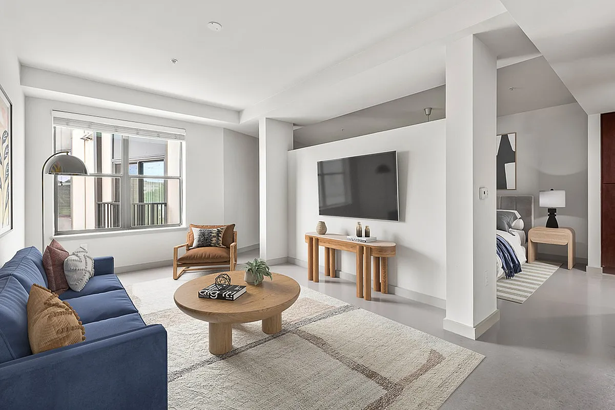 Modern living room at Cottonwood Westside Apartments with a blue sofa, wooden furniture, large window, and wall-mounted TV, leading to a cozy bedroom.