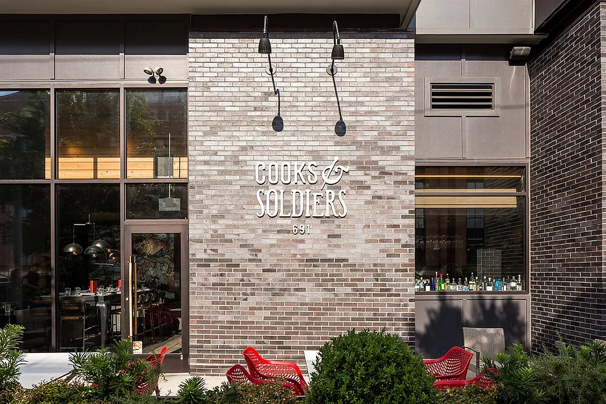 Charming brick facade of Cooks & Soldiers with stylish red chairs and lush plants, evoking a welcoming and vibrant atmosphere.