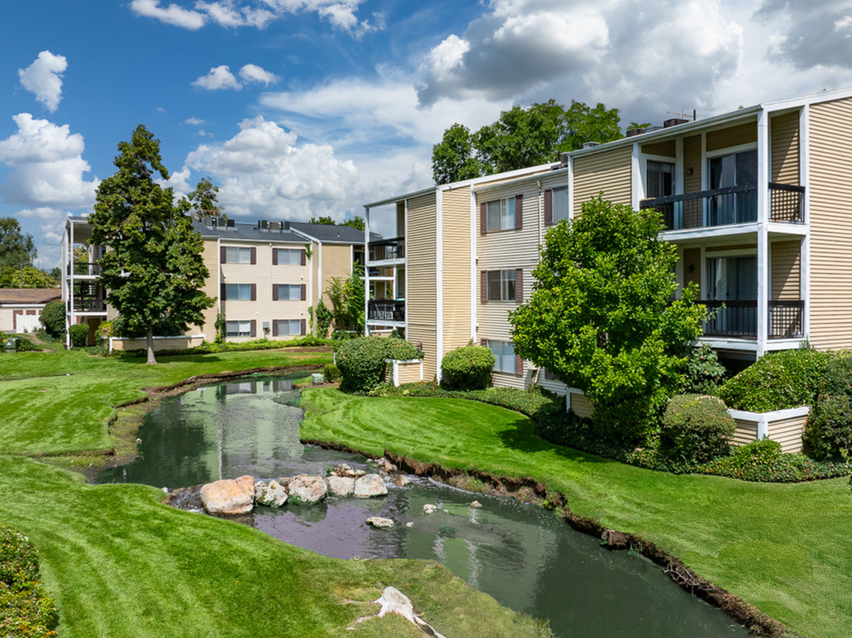 Charming apartment complex, Fox Point in Old Farm, with serene landscaped gardens, a tranquil stream, and vibrant greenery under a bright blue sky.