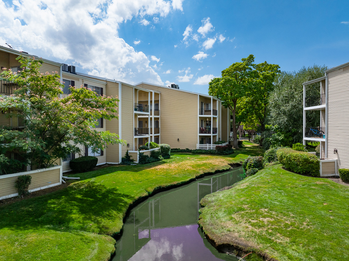 Charming apartment complex, Fox Point in Old Farm Apartments, with picturesque stream and lush landscaping under a bright, blue sky.