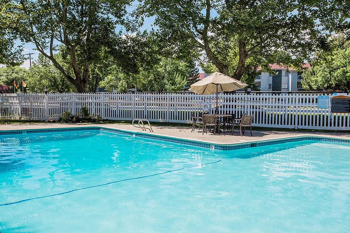 Charming outdoor pool area Fox Point in Old Farm Apartments with sparkling water, surrounded by lush greenery and a cozy seating area under an umbrella.