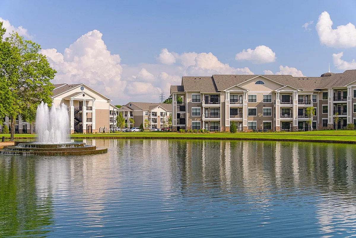Luxurious apartment complex, Heights at Meridian, overlooking a serene pond with a fountain, surrounded by lush greenery under a bright blue sky.