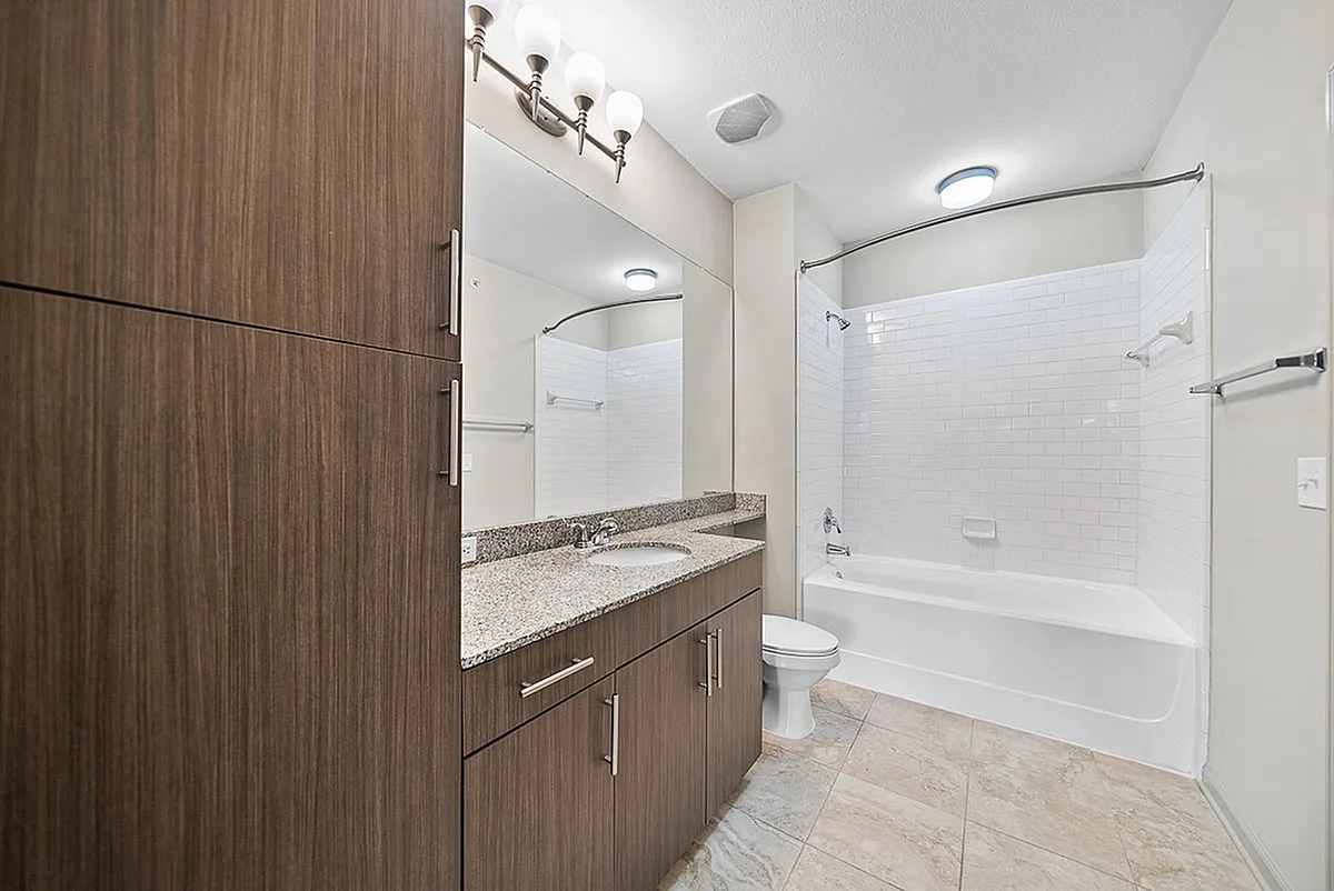 Elegant bathroom at Heights at Meridian Apartments with modern wooden cabinetry, granite countertops, and a sleek tub surrounded by white subway tiles.