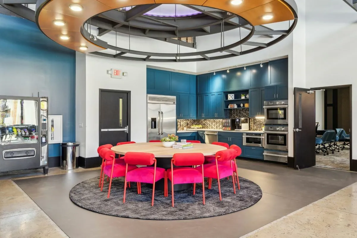 Modern communal kitchen with vibrant red chairs around a circular table, surrounded by blue cabinets and equipped with stainless steel appliances, perfect for social gatherings at The Melrose in Nashville.