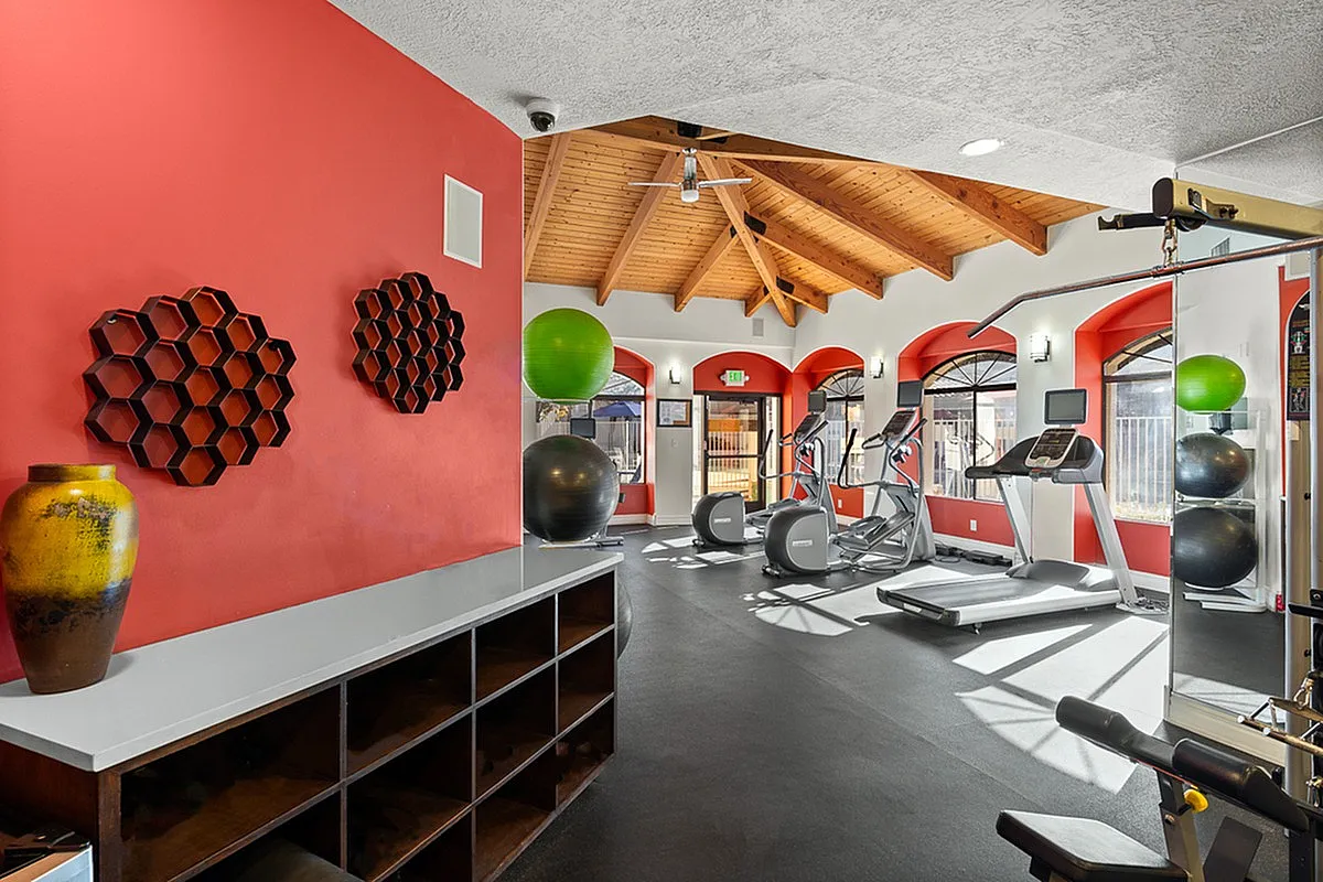 Bright and inviting fitness room featuring state-of-the-art exercise equipment, vibrant red accent wall, and large windows for natural light at Pavilions Apartments.