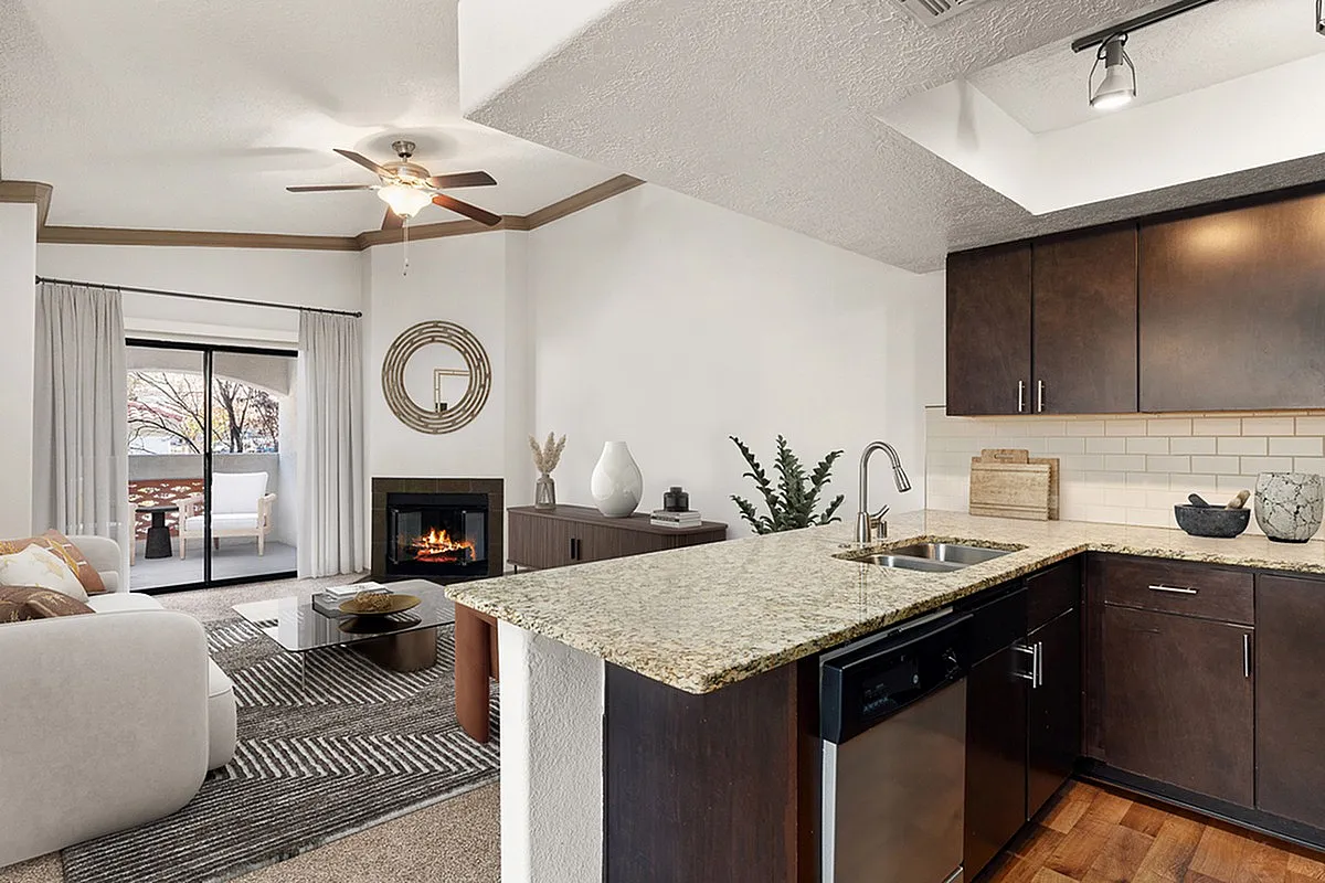 Contemporary open-plan living space at Pavilions Apartments featuring elegant kitchen with dark cabinets, cozy living room with fireplace, and private balcony view.