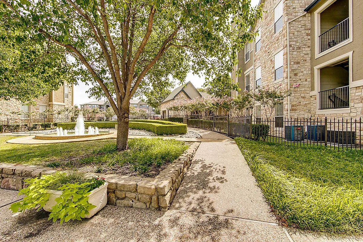 Stonebriar of Frisco's charming apartment exterior with a serene pathway, lush greenery, and a sparkling fountain, perfect for nature lovers.