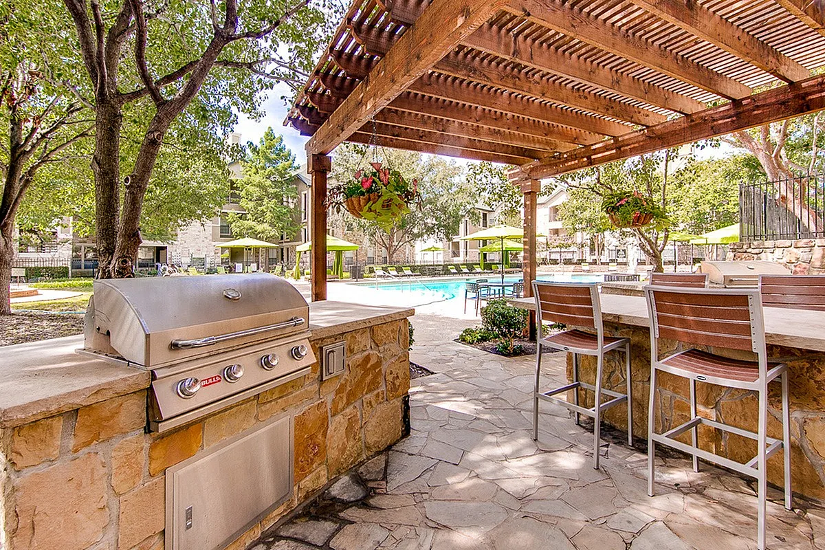Charming outdoor area featuring a spacious grill under a wooden pergola, with a beautifully landscaped poolside view and comfortable seating at Stonebriar of Frisco Apartments.