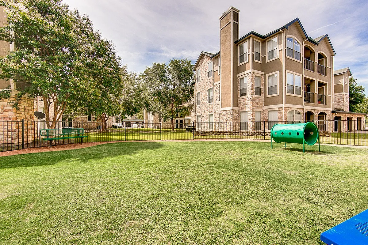 Charming apartment complex, Stonebriar of Frisco Apartments, featuring elegant architecture with lush green lawns and a cozy play area for relaxation.