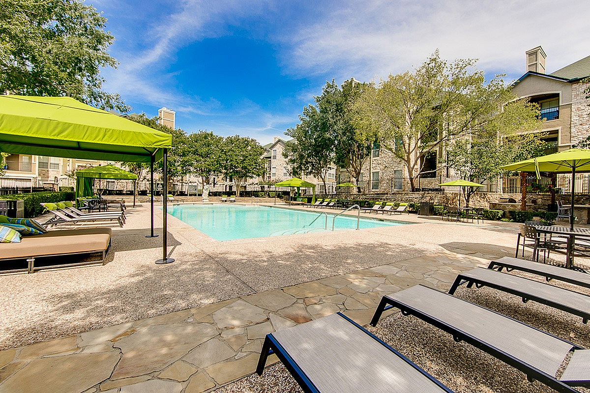 Inviting outdoor pool area at Stonebriar of Frisco Apartments with elegant lounge chairs and vibrant green canopies, nestled amidst lush greenery and modern apartment buildings.