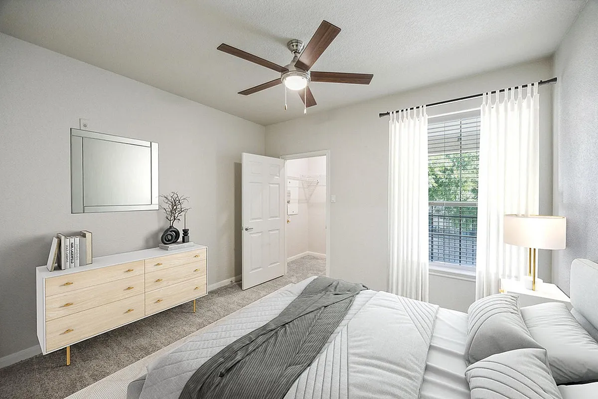 Bright and inviting bedroom at Stonebriar of Frisco Apartments with modern decor featuring a stylish dresser, ceiling fan, and large window with sheer curtains.