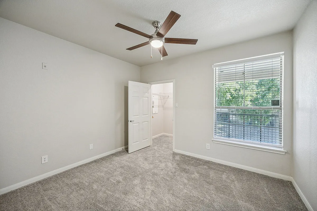 Bright bedroom with modern ceiling fan, plush carpet, and large window offering green views at Stonebriar of Frisco Apartments.